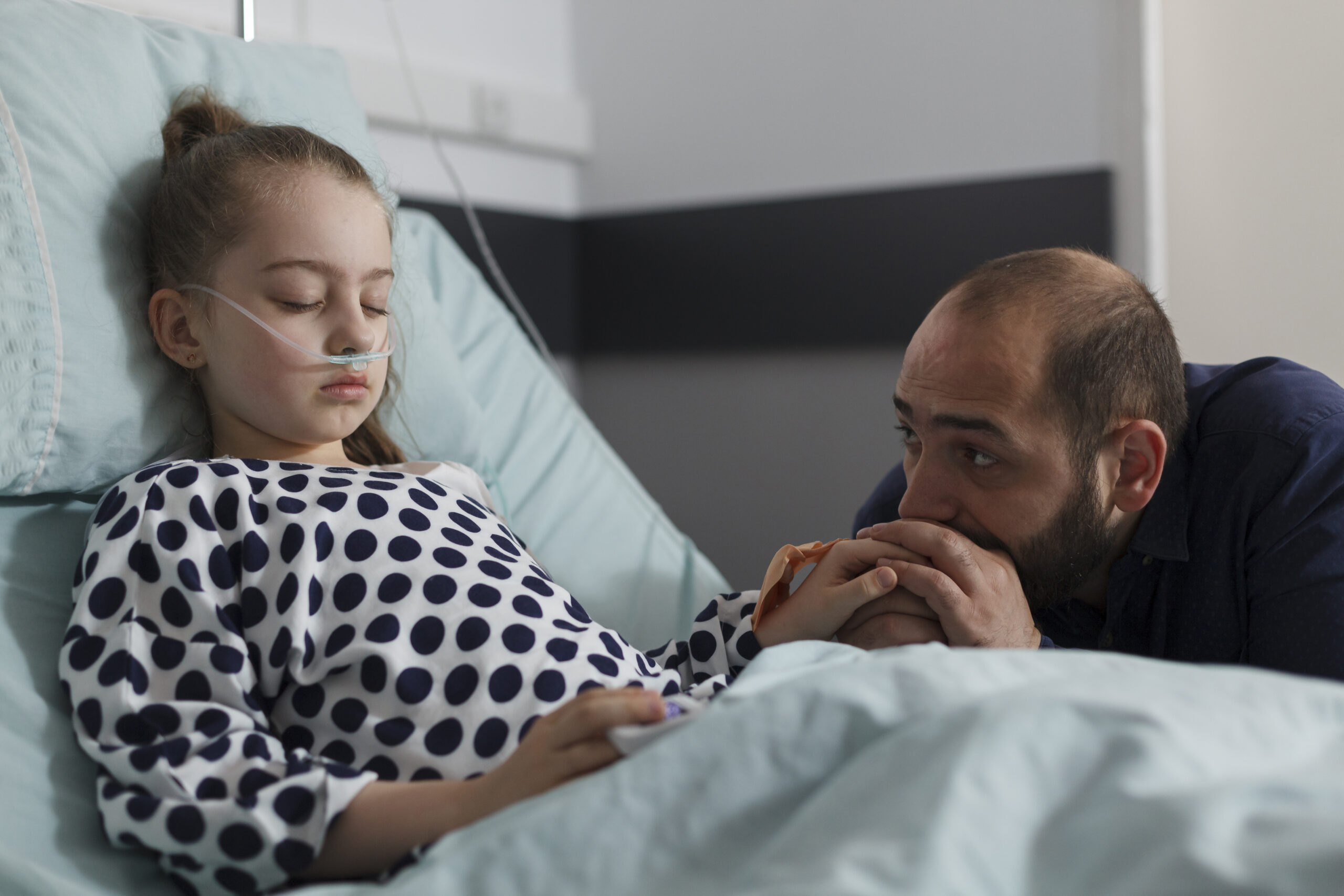 Unwell sick kid sleeping while uneasy parent sitting beside her inside healthcare facility. Worried father sitting next to ill little girl resting on patient bed inside hospital pediatrics ward room.